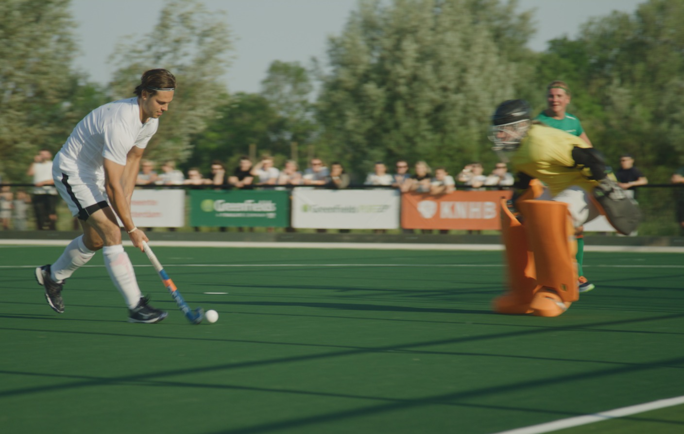 Field hockey player in white dribbling the ball on an artificial turf pitch, approaching a goalkeeper in protective gear.