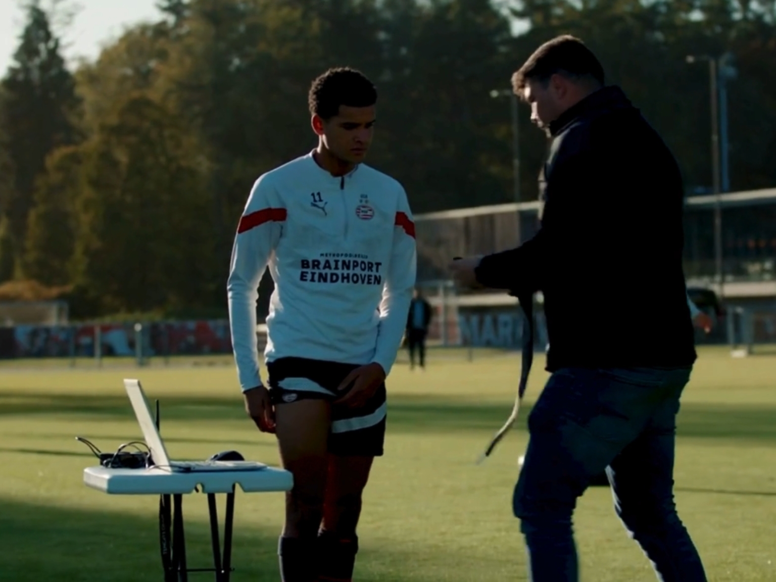 Soccer player on a training field being fitted with a monitoring strap by a staff member, with a laptop set up on a table nearby.