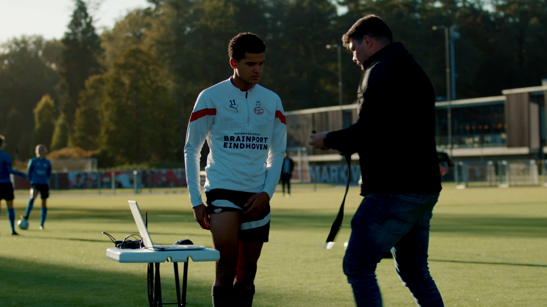 Soccer player on a training field being equipped with a monitoring strap by a staff member, with a laptop set up nearby.