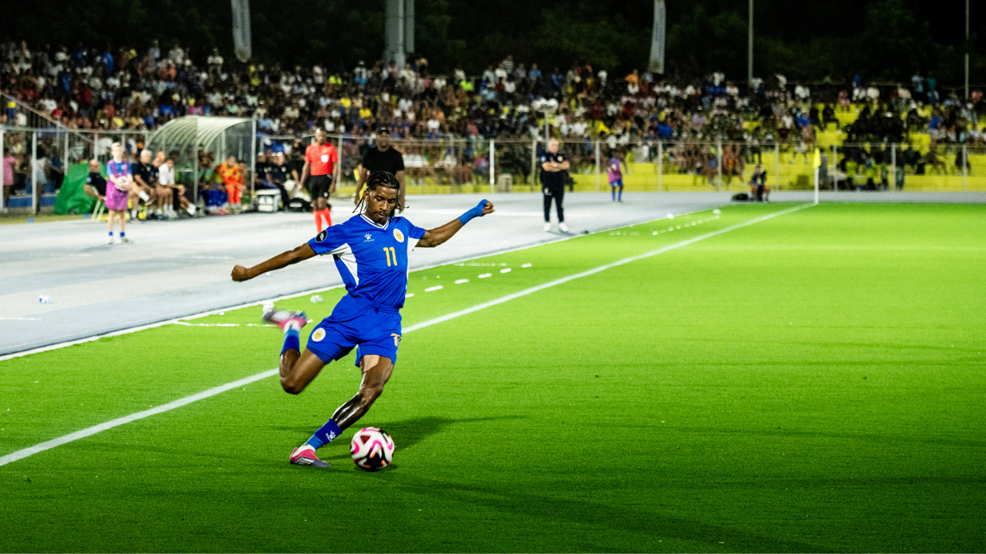 Soccer player in a blue uniform kicking the ball on an artificial turf field during a night match, with a large crowd in the stands.