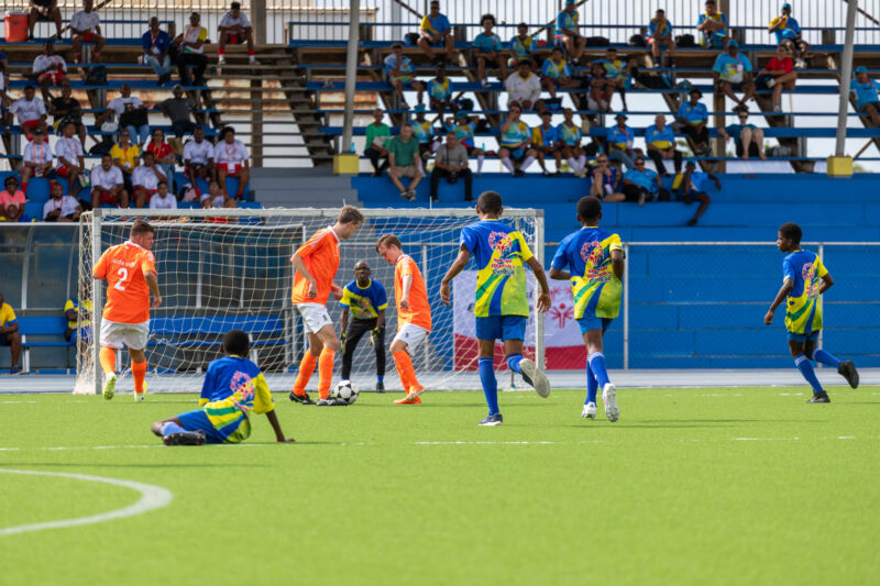 Youth soccer match on an artificial turf field, with players in orange and blue uniforms competing near the goal while spectators watch from the stands.